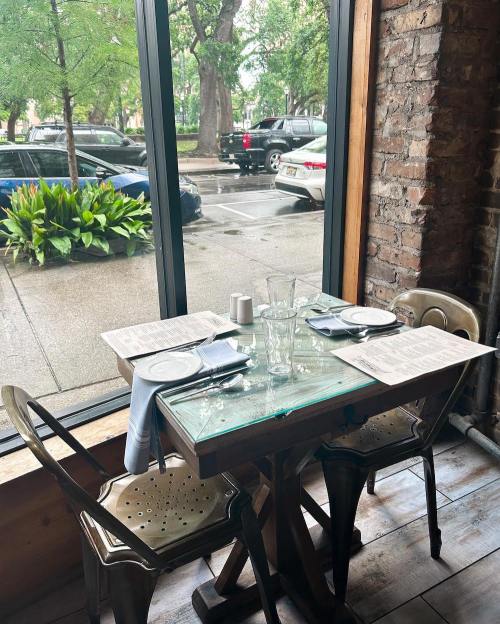 A cozy restaurant table set for two by a large window, overlooking a tree-lined street.
