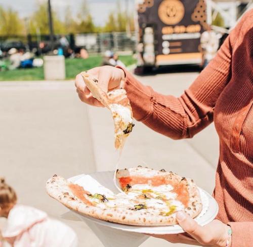 A person holds a slice of pizza with melted cheese, topped with herbs, over a plate in an outdoor setting.