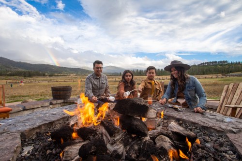 Four friends gather around a fire pit, roasting marshmallows with a scenic landscape and rainbow in the background.