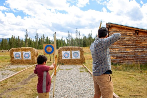 A man and a boy practice archery at an outdoor range with targets set up on hay bales in a scenic landscape.