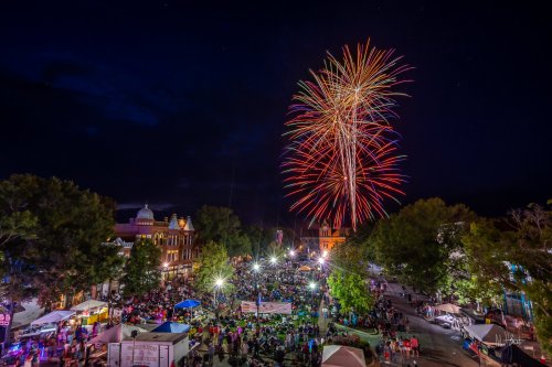 Colorful fireworks illuminate the night sky over a crowded outdoor event, with trees and historic buildings in the background.