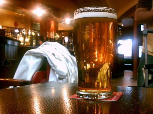 A glass of amber beer sits on a wooden table in a cozy pub, with a blurred background of bar shelves.
