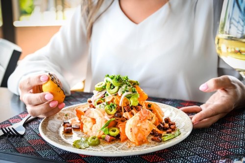 A person holds a lemon slice while enjoying a plate of shrimp pasta topped with greens and olives.