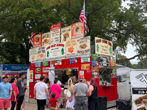 A busy food stand at a fair, featuring colorful signs and a crowd of people waiting in line.