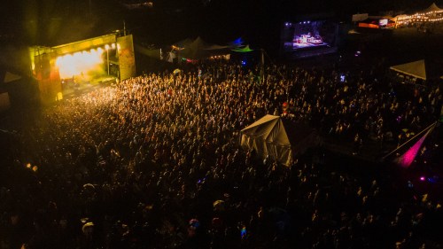 Aerial view of a large crowd at a music festival, with a stage illuminated by bright lights in the background.