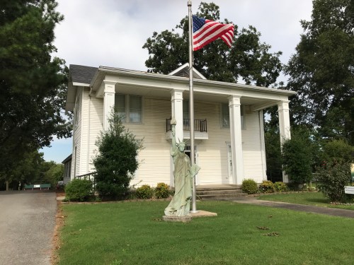 A white building with a porch, featuring a statue of the Statue of Liberty and an American flag in front.