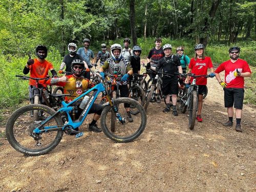 A group of mountain bikers poses together on a trail, surrounded by trees and dirt paths.