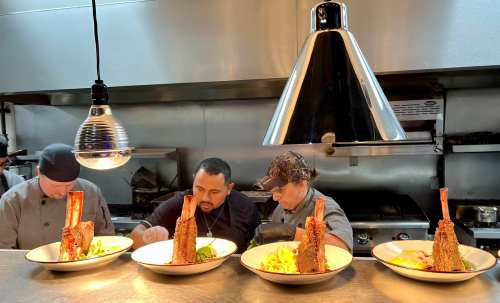 Three chefs in a kitchen prepare dishes with large meat cuts on plates, under a hanging light fixture.