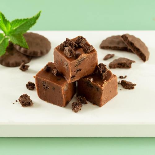 Three chocolate fudge squares topped with cookie crumbles, surrounded by mint leaves and broken chocolate cookies on a white plate.