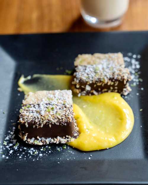 Two chocolate squares topped with coconut and pistachios, served on a black plate with a yellow sauce.