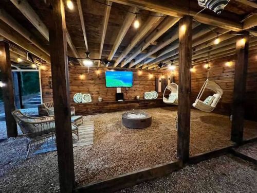 Cozy outdoor lounge with hanging chairs, a TV, and a fire pit, surrounded by wooden beams and warm lighting.