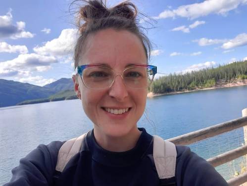 A smiling person with glasses stands by a lake, surrounded by mountains and trees under a blue sky with clouds.