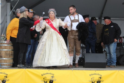 A woman in a gown and crown stands on stage with a man in traditional attire, both engaging with the audience at a festival.