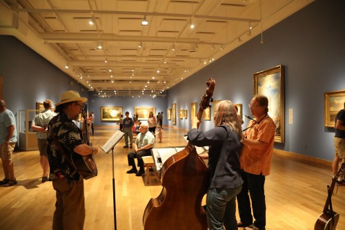 Musicians perform in an art gallery while visitors admire paintings on the walls.