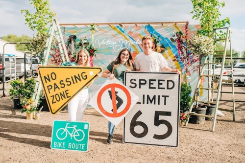 Three people hold various road signs, including "No Passing Zone," "Speed Limit 65," and "Bike Route," in a colorful setting.