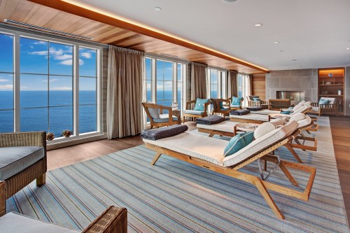 Bright, modern lounge with wooden furniture, large windows overlooking the ocean, and a cozy atmosphere.
