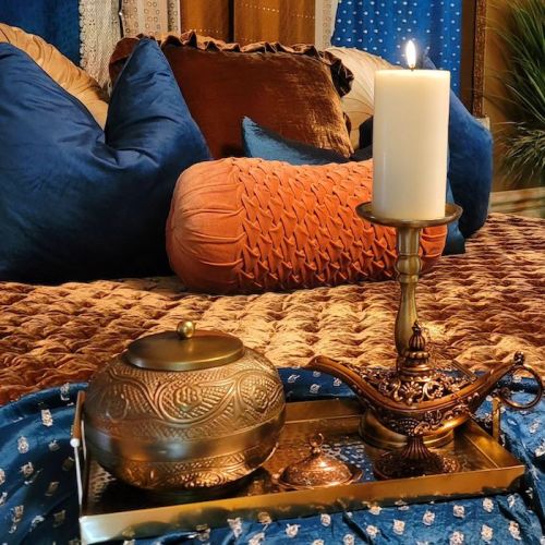 A cozy setting with a lit candle, decorative bowl, and ornate lamp on a tray, surrounded by plush pillows and fabrics.