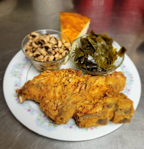 A plate featuring fried fish, black-eyed peas, collard greens, and a slice of cornbread on a floral-patterned plate.
