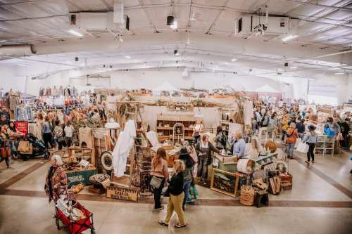 A bustling indoor market filled with vendors, shoppers, and various vintage items displayed throughout the space.