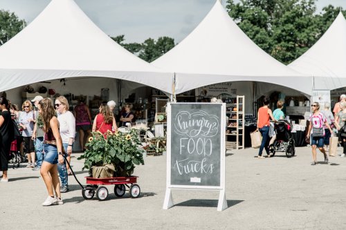 A bustling outdoor market with tents, people shopping, and a sign for food trucks. A woman pulls a wagon with plants.