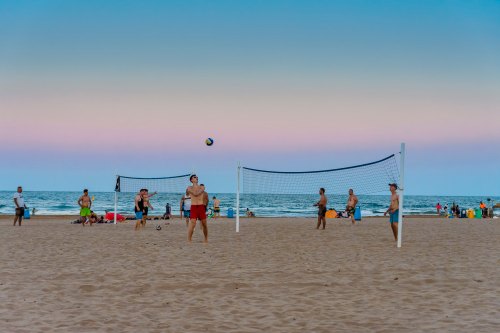 People playing beach volleyball on a sandy shore with the ocean and a colorful sky in the background.