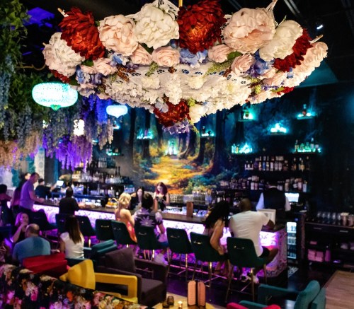 A vibrant bar scene with a floral chandelier, colorful seating, and patrons enjoying drinks in a lively atmosphere.