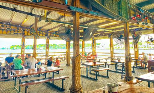 A lively waterfront restaurant with wooden tables, fans, and diners enjoying the view.