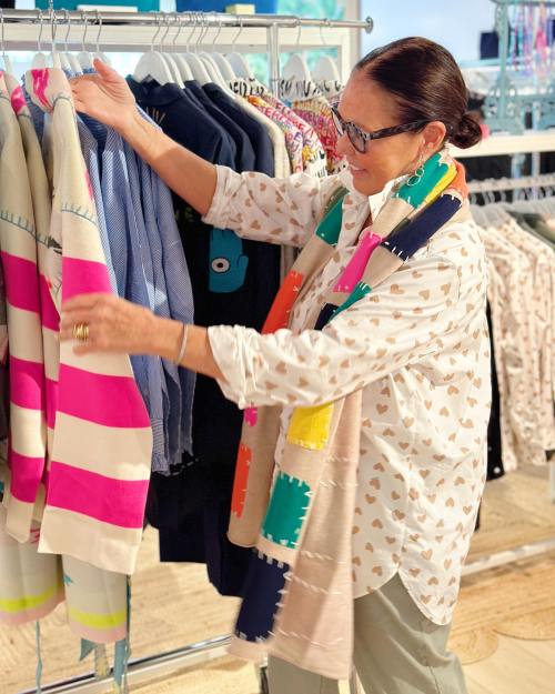 A woman examines colorful clothing on a rack in a bright store, wearing a patterned scarf and a white shirt.