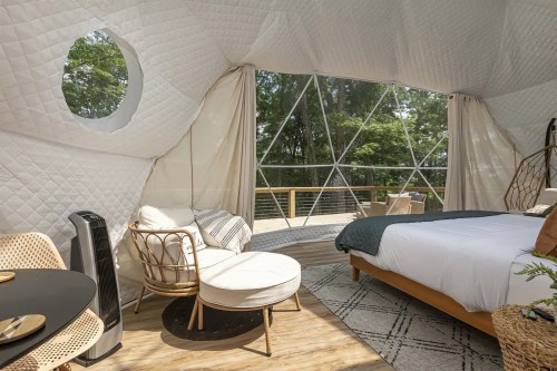 Cozy geodesic dome interior with a bed, seating area, and large windows overlooking a forested view.