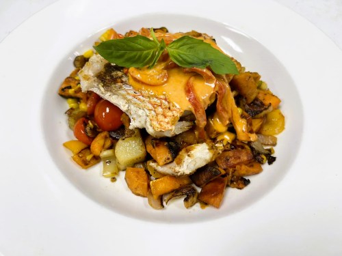 A plate of sautéed vegetables topped with a piece of fish, garnished with basil and a drizzle of sauce.