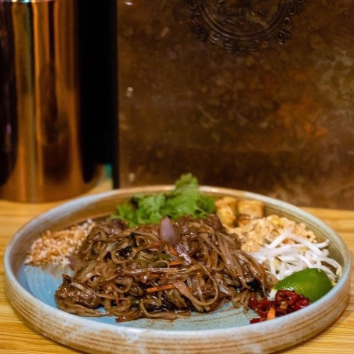 A plate of noodles with vegetables, herbs, and lime, served with toppings on a wooden table.