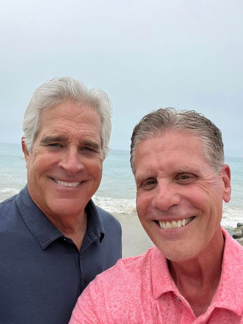 Two smiling men pose for a selfie by the beach, with a cloudy sky and ocean in the background.