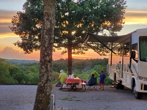 A family enjoys a sunset picnic by their RV under a tree, surrounded by lush greenery and a colorful sky.