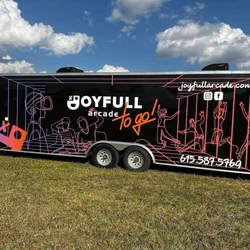 A black trailer with colorful graphics promoting "Joyfull Arcade to Go!" against a blue sky and grassy field.