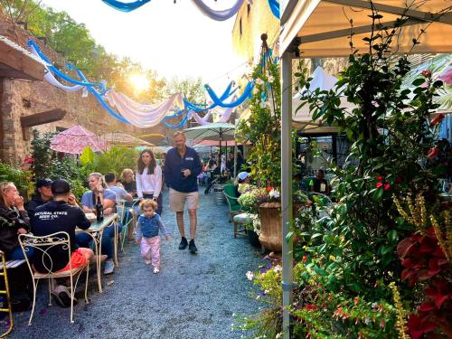 A lively outdoor dining area with people, colorful umbrellas, and lush greenery, illuminated by warm sunlight.