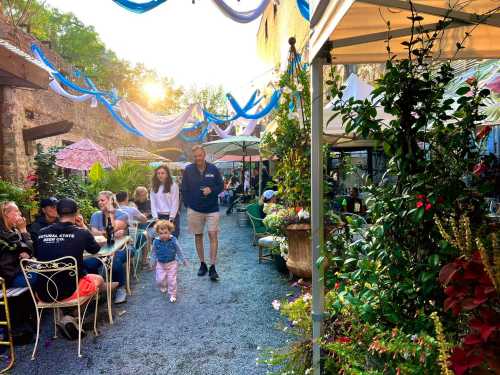 A lively outdoor dining area with people, colorful umbrellas, and lush greenery, illuminated by warm sunlight.