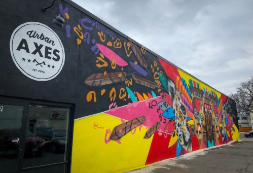 Colorful mural on a building wall featuring abstract designs and shapes, with the "Urban Axes" logo visible.