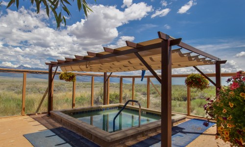 A serene outdoor hot tub under a shaded pergola, surrounded by lush greenery and a scenic sky with fluffy clouds.