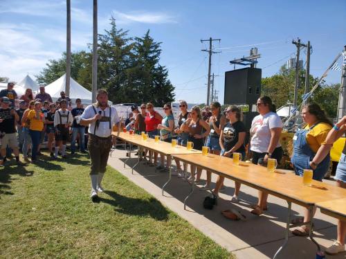A crowd watches a man in lederhosen at a beer-drinking contest, with participants lined up at a long table outdoors.