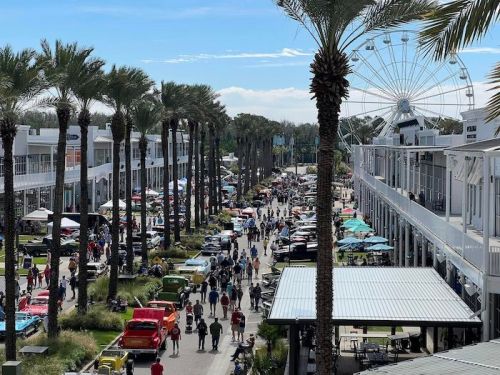 A bustling outdoor market with palm trees, classic cars, and a Ferris wheel in the background on a sunny day.