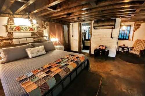 Cozy bedroom with a metal bed, colorful quilt, wooden beams, and rustic decor, featuring a small sitting area.