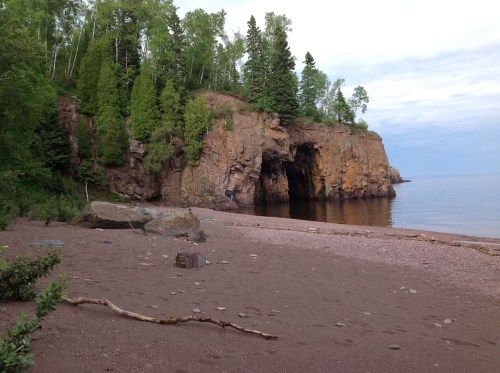 A serene beach scene with a rocky cliff, lush trees, and calm water under a cloudy sky.