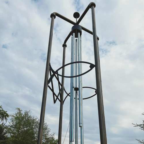 A tall, modern sculpture with metal tubes and circular elements against a cloudy sky.