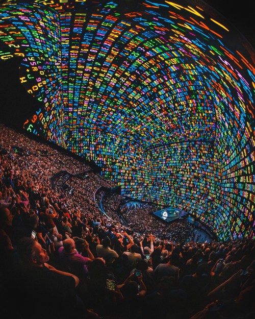A vibrant, colorful digital display illuminates a packed auditorium, showcasing a lively event with an engaged audience.