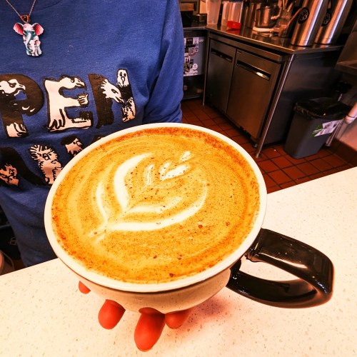 A person holding a large cup of latte art, wearing a blue shirt with a playful design in a café setting.