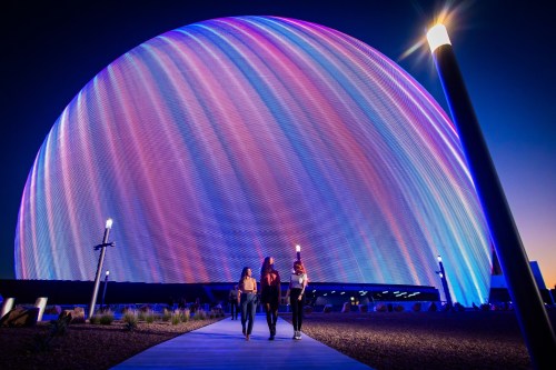 Three people walk along a path in front of a large, illuminated dome structure at dusk.