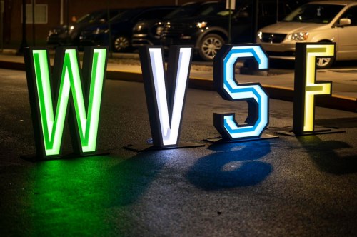 Brightly lit letters "W," "V," "S," and "F" stand on a dark street, with parked cars in the background.