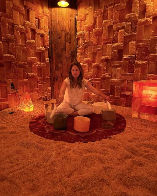 A woman sits on a rug in a salt room, playing crystal singing bowls surrounded by salt brick walls and soft lighting.