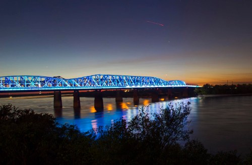 A brightly lit blue bridge spans a river at dusk, with a colorful sunset in the background.