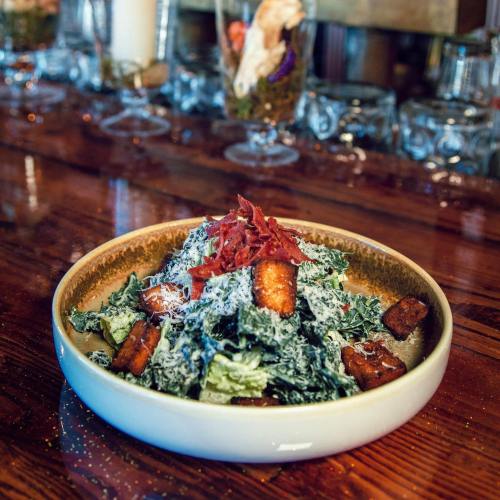 A bowl of salad with greens, croutons, cheese, and crispy toppings, placed on a wooden table with a bar in the background.